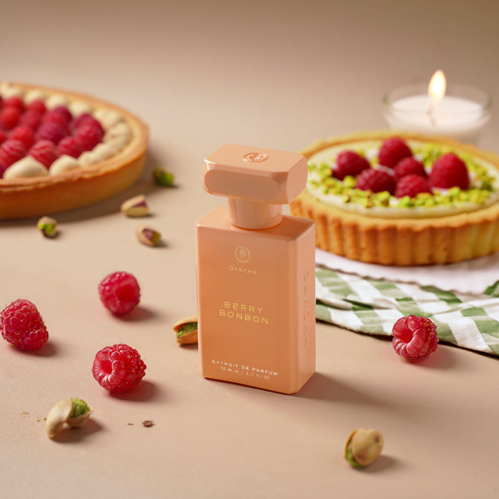 Front view of Berry Bonbon perfume, peach pink rectangular bottle with gold cap beside raspberries and tarts on a beige surface