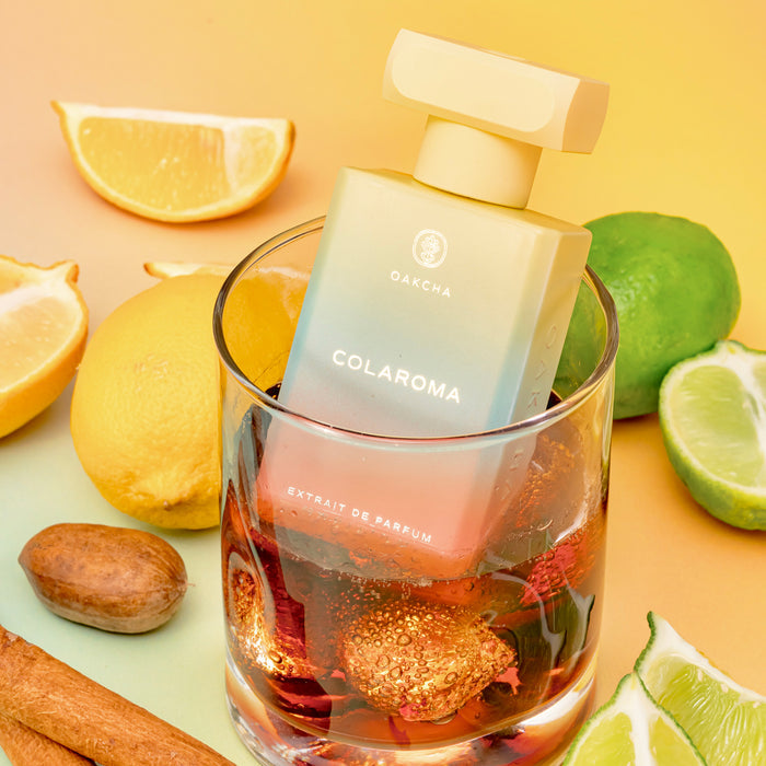Angled front view of Colaroma perfume bottle with gradient finish in a glass of amber liquid, surrounded by lemon and lime