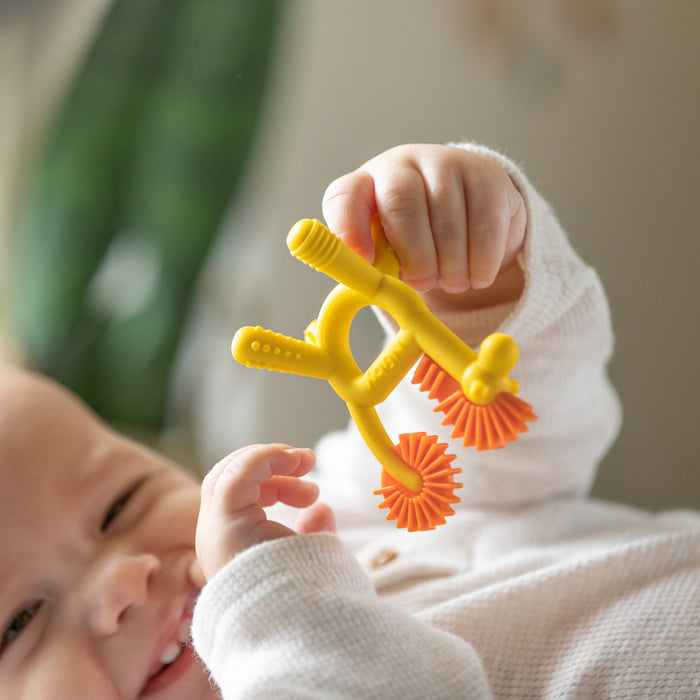 A smiling baby in white holds up a yellow and orange Nuby Geo Zoo Silicone Teether while lying down, with a blurred green plant in the background.