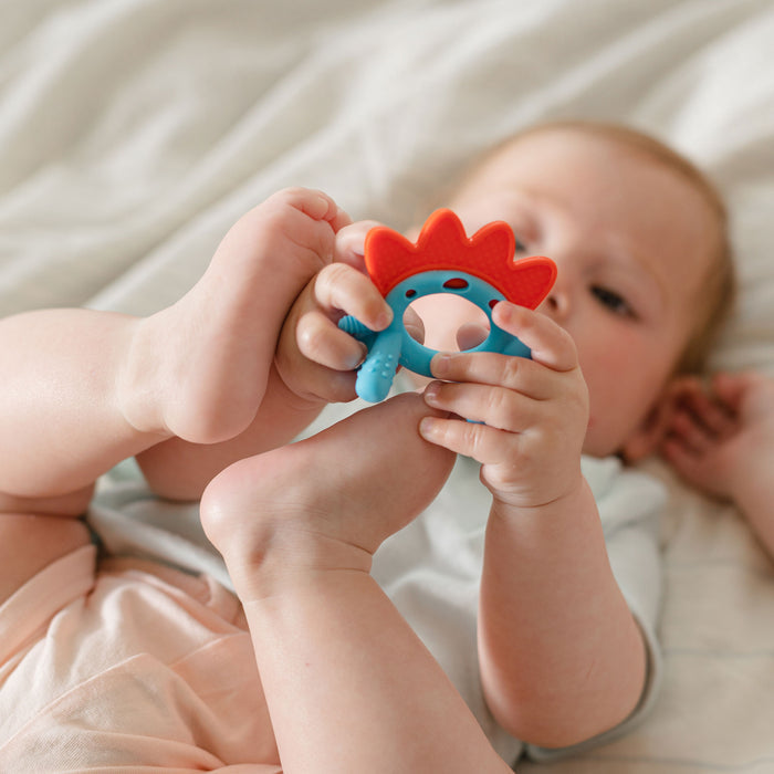 A baby lies on a bed, holding the Nuby Geo Zoo Silicone Teether with both hands and feet, and looks at it closely.
