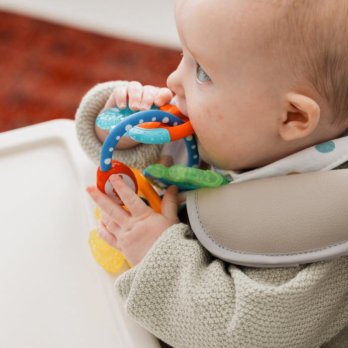 A baby in a high chair chews on IcyBite Teether Keys | Classic, holding them with both hands. Dressed in a light sweater and pastel dot bib, the baby enjoys soothing teething relief.
