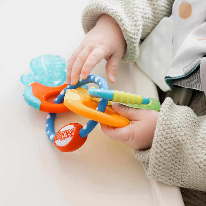 A baby in a gray sweater holds the IcyBite Teether Keys | Classic with orange, blue, green, and translucent parts while sitting at a white surface—perfect for teething relief.