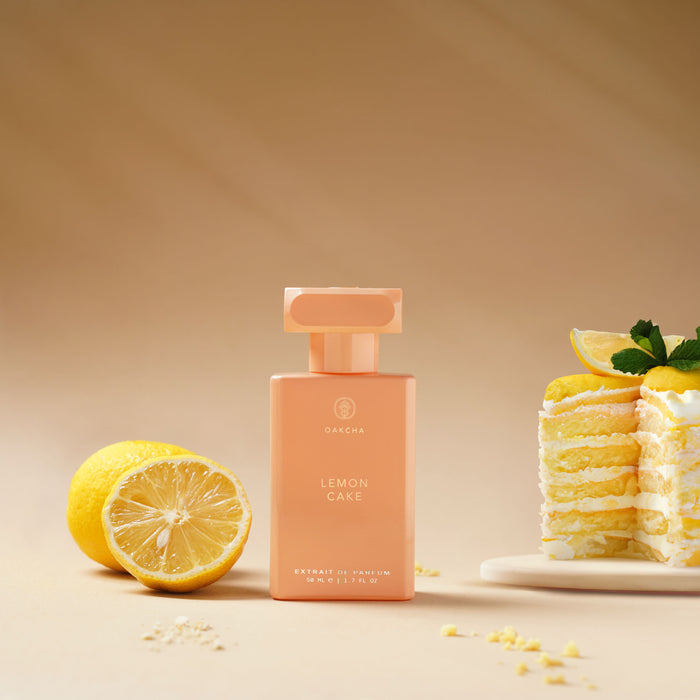 Front view of Lemon Cake perfume bottle in peach-pink glass with gold cap beside half a lemon and a pancake stack on warm background
