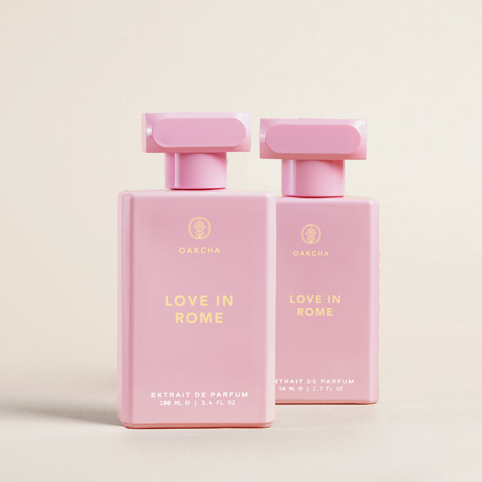 Front view of two pink Love In Rome perfume bottles showcasing rectangular caps, gold Oakcha logo and LOVE IN ROME text on a pale beige background