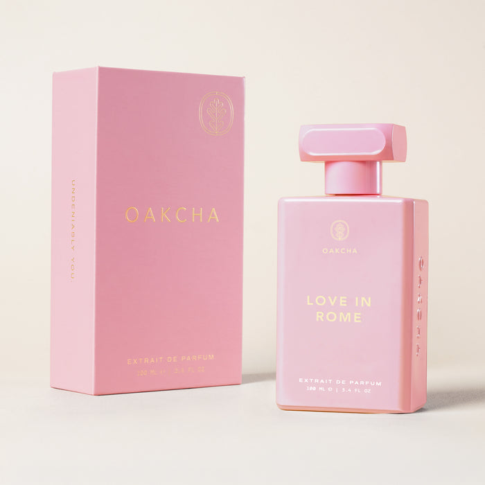 Front view of Love in Rome 100 mL perfume bottle and box, pink bottle with gold lettering on a light background
