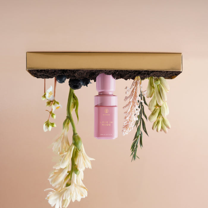 Front view of Love in Rome perfume oil bottle hanging upside down under a gold bar with white flowers on a beige background