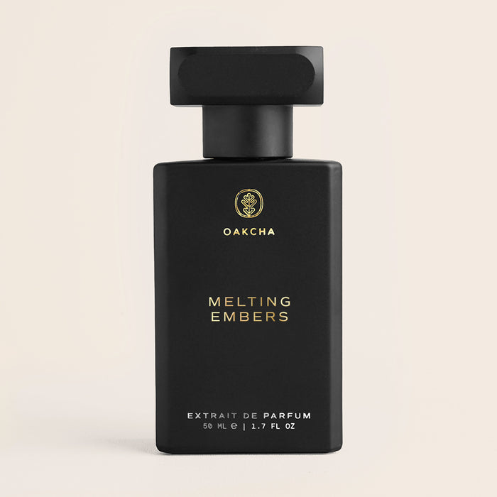 Front view of Melting Embers perfume bottle with matte black rectangular design, gold logo and text, and black cap on beige background