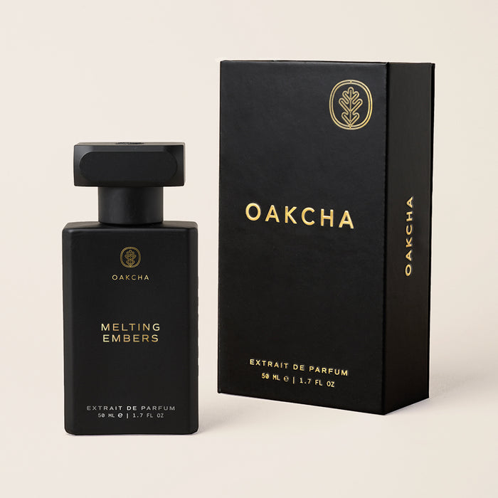 Front view of Melting Embers fragrance bottle and box, matte black rectangular bottle with gold logo on beige background