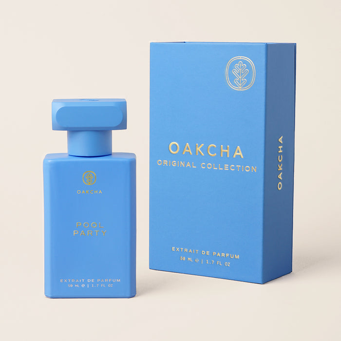 Front view of Pool Party perfume bottle and matching box showcasing matte sky-blue bottle with gold logo on beige studio background