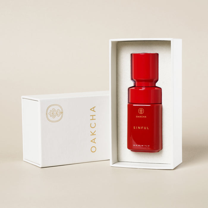Front view of Sinful Perfume Oil showcasing glossy red bottle nestled in white presentation box against a beige background