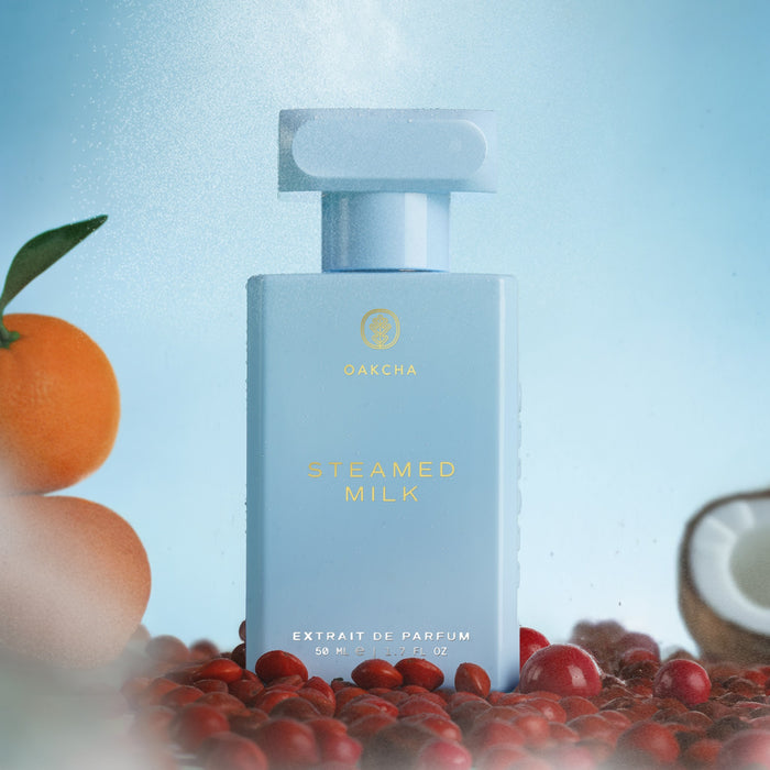 Front view of Steamed Milk perfume bottle, pale blue with gold logo, on blue background with orange, coconut, and red berries