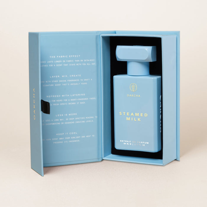 Front view of Steamed Milk bottle showcasing rectangular light-blue bottle and gold cap inside an open matching box on a beige background