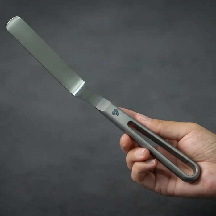 Hand holding a offset spatula with a gray handle and blade against a dark background