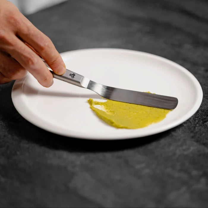 Hand holding a offset spatula with a gray handle and blade spreading a yellow condiment on white plate against a dark background.