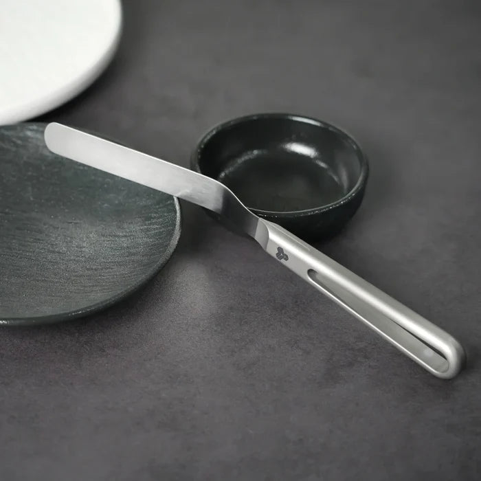 An offset spatula with a gray handle and blade against laying on a dark grey plate with black bowl behind it on dark grey background.